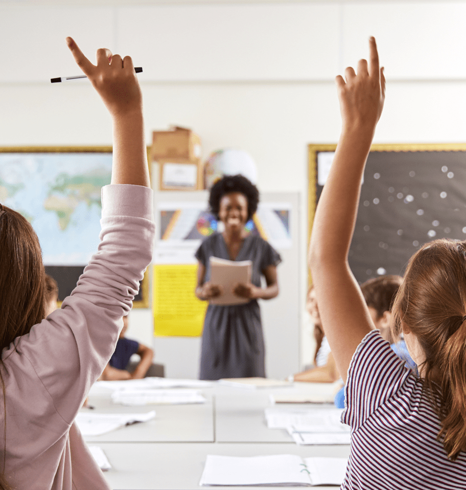 Élèves levant la main dans la salle de classe. L’enseignante sourit en arrière-plan.