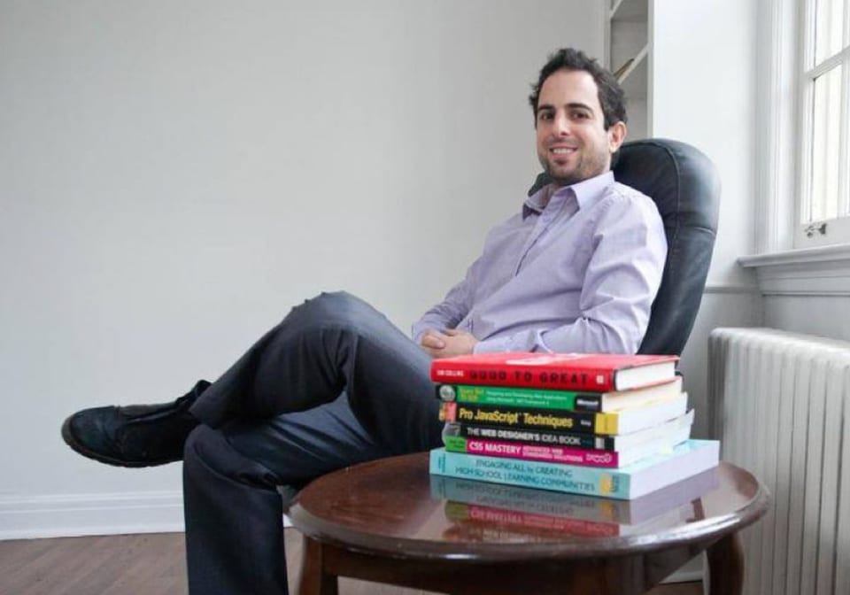 Gil Silberstein, smiling, sitting on a chair next to a stack of coding books on a table.